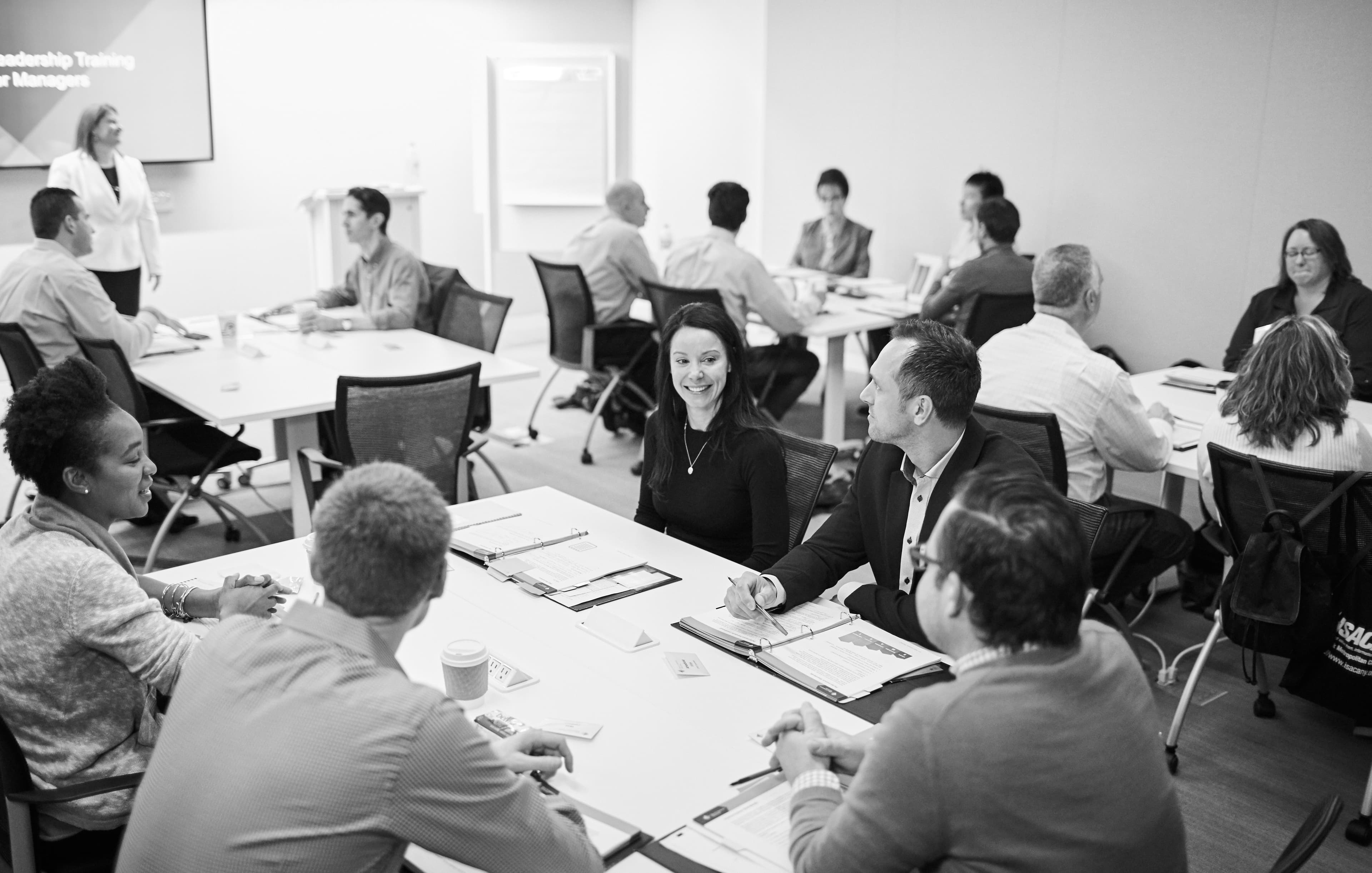 Turma reunida em um treinamento de liderança Dale Carnegie, com facilitadora conduzindo a sessão ao fundo
